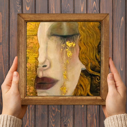 Framed artwork of a woman's face with gold accents held by hands against a wooden background