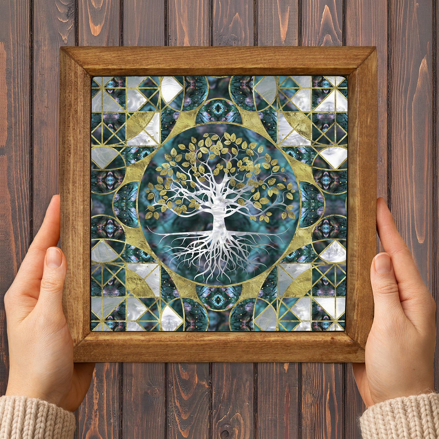 Framed artwork with a tree design held by two hands on a wooden background