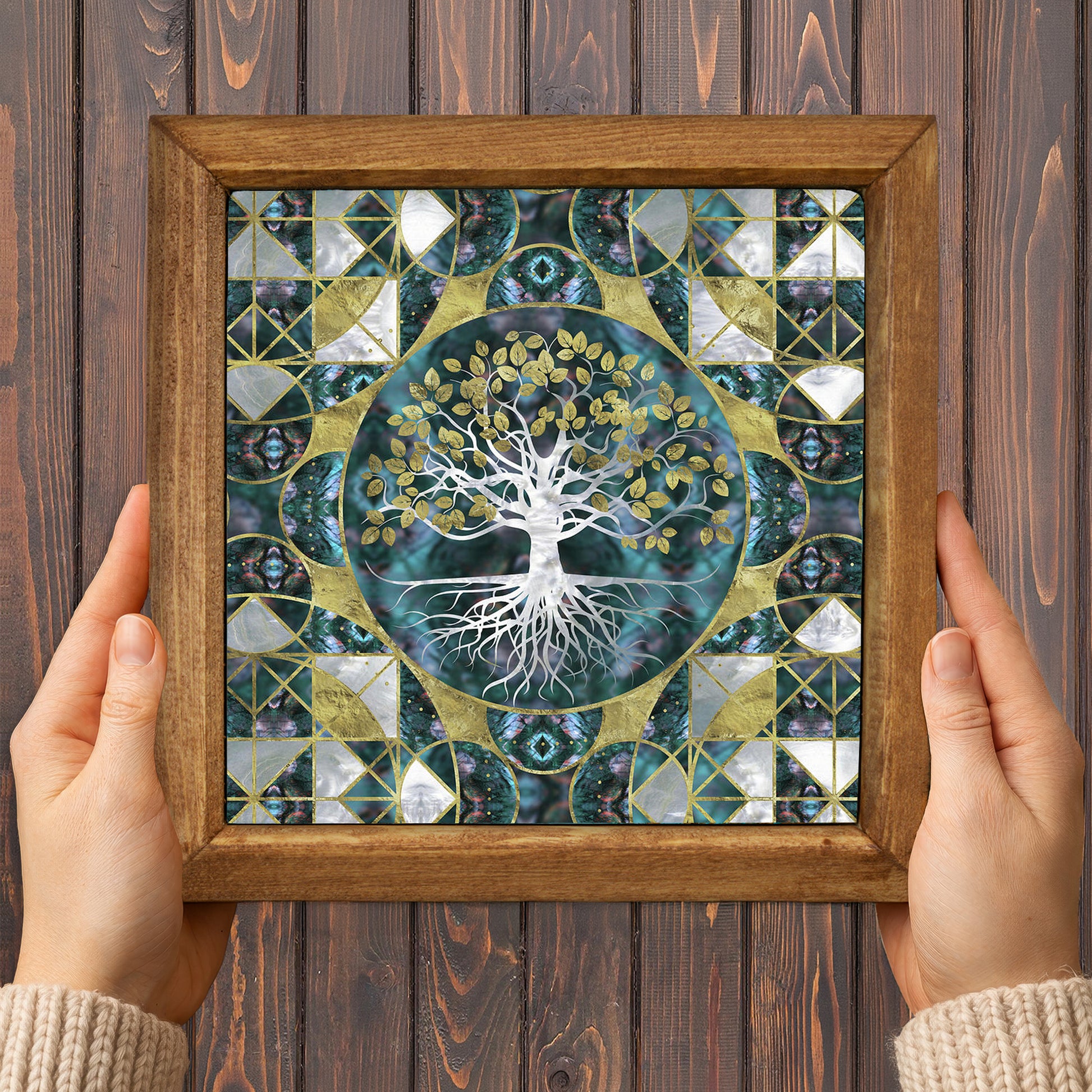 Framed artwork with a tree design held by two hands on a wooden background