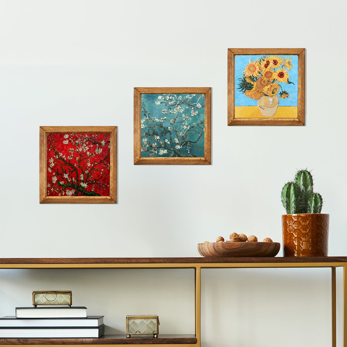 Three framed paintings on a wall above a wooden shelf with decorative items.